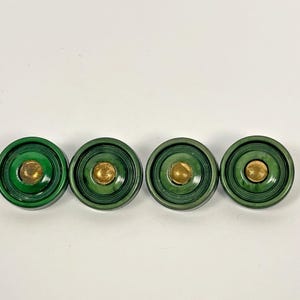 May include: Four green, circular cabinet knobs with a brass center. The knobs have a layered design, with a darker green outer ring and a lighter green inner ring. The brass center adds a touch of elegance to the design.
