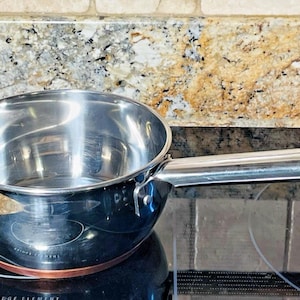 May include: A stainless steel saucepan with a long handle rests on a black electric stovetop. The pan has a shiny, reflective surface and a copper-colored base. The background features a speckled granite countertop.