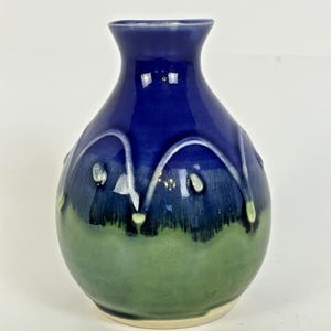 May include: A small, handcrafted ceramic vase with a glossy finish. The upper portion is a deep blue, transitioning to a green base. White, curved accents adorn the blue section. The vase has a rounded body and a narrow neck.