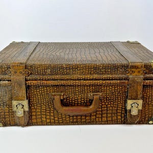 May include: A vintage brown suitcase with a textured, crocodile-like pattern. The suitcase features brass corner protectors, leather straps, and a handle. The clasps are ornate, and the overall design suggests a classic travel accessory.