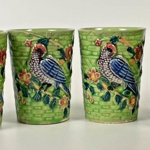May include: A set of four cylindrical ceramic tumblers featuring a raised relief design of a blue and grey parrot perched on a branch. The background has a light green basket-weave texture with small pink flowers and green leaves throughout.