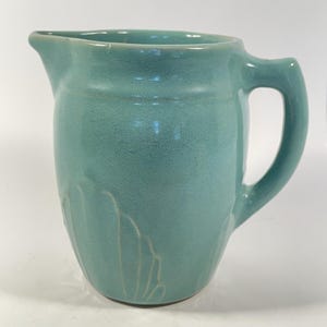 May include: A turquoise ceramic pitcher with a curved handle and a spout. The pitcher has a textured surface and a decorative, embossed design on the lower portion of the body. The pitcher is designed for serving beverages.