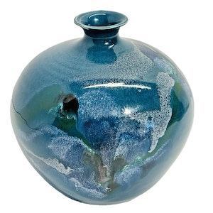 May include: A teal-colored ceramic vase with a round body and a narrow neck. The vase has a glossy finish with streaks of white and darker blue, creating a marbled effect. A dark hole is visible on the side of the vase.
