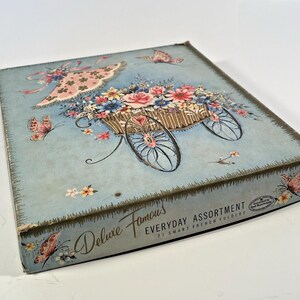 May include: A vintage box with a light blue background and a floral design. The box features a decorative cart filled with flowers, butterflies, and the text "Deluxe Famous Everyday Assortment". The box has a distressed, aged appearance.