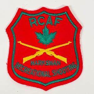 May include: A shield-shaped embroidered patch with a red background and green border. It features a green maple leaf, two crossed yellow rifles, and the text RCAF, MARKSMAN, and RECREATIONAL SHOOTING in green embroidery on the red fabric surface.