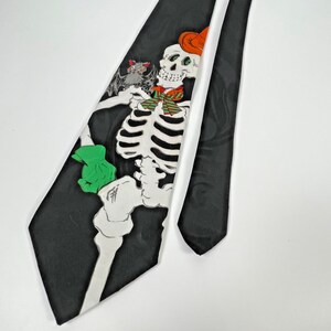 May include: A black necktie featuring a white skeleton graphic wearing an orange hat and a green and red striped bowtie. The skeleton holds a small grey bat in one hand and wears a bright green glove on the other hand, set against a black background.