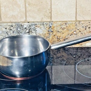 May include: A stainless steel saucepan with a long handle. The pan is sitting on a black electric stovetop with a copper-colored heating element. The background features a granite countertop and beige tile backsplash.