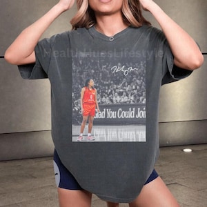 May include: Dark gray t-shirt featuring a black and white image of a basketball player in a red uniform. The image includes the text "Glad You Could Join" and a signature. The shirt has a V-neck.