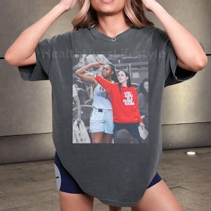 May include: Dark gray t-shirt with a printed image of two women on a basketball court. One woman is wearing a red sweatshirt with text, and the other is in a basketball uniform. The shirt has a v-neck and short sleeves.