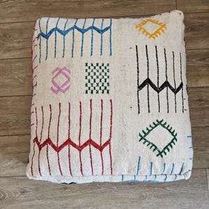 Peut inclure: Pouf carré blanc cassé avec un motif géométrique bleu, rouge, jaune, vert et noir. Le motif comprend des zigzags, des lignes verticales et des losanges. Le pouf est posé sur un sol en bois.