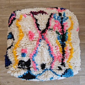Peut inclure: Un pouf blanc et moelleux avec un motif géométrique coloré. Le motif comprend du rose, du bleu, du jaune, du noir et du vert. Le pouf est de style marocain traditionnel.