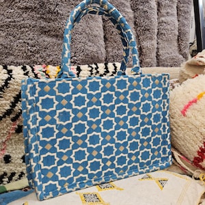 May include: A blue tote bag with a geometric pattern of white and gold shapes. The bag has matching handles and a white border. The bag is sitting on a patterned rug. The text "Tifinagh Shop" is visible.