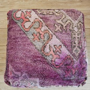 Peut inclure: Un pouf carré, de couleur violet et rose, avec un motif géométrique. Le pouf est fait d'un matériau doux et duveteux.