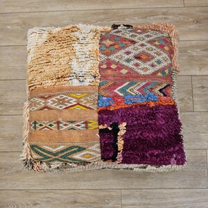 Peut inclure: Un coussin de sol carré avec un design patchwork. Il présente des sections en tissu shaggy beige et violet, ainsi que des panneaux à motifs rouges, oranges, bleus et verts. Un élément décoratif pour la maison avec diverses textures.