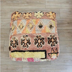 Peut inclure: Un pouf marocain coloré avec un motif géométrique dans des tons de rose, orange, jaune et marron. Le pouf est fait de tissu tissé et a une texture touffetée.