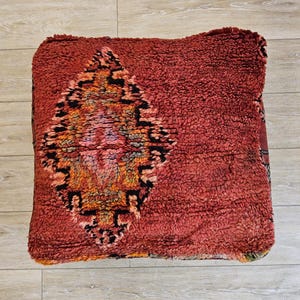 Peut inclure: Un pouf carré rouge texturé avec un motif géométrique central noir, orange et jaune. Le pouf a un aspect moelleux et invitant, suggérant confort et une esthétique bohème. Le logo Tifinagh Shop est visible.