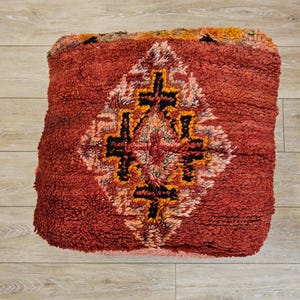 Peut inclure: Un pouf carré rouge texturé avec un motif central en losange noir, orange et crème. Le pouf a un aspect hirsute et tissé, suggérant un style fait main ou vintage. Le design est géométrique et ethnique.