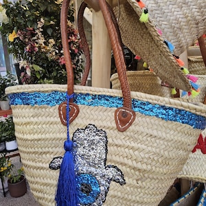 May include: Woven straw tote bag with brown leather handles and a blue sequin trim. The bag features a sequined Hamsa hand design and a blue tassel. Additional straw hats with colourful tassels are visible in the background.