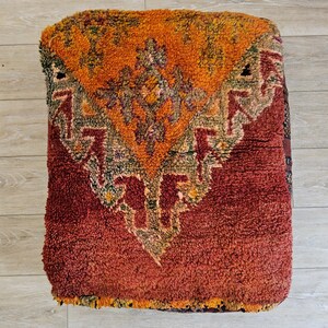 Peut inclure: Un pouf carré et texturé dans les tons orange et rouge. Le dessus présente un motif géométrique central en orange, vert et rouge. Le pouf est fait d'une matière épaisse et tissée, et repose sur un sol en bois clair.