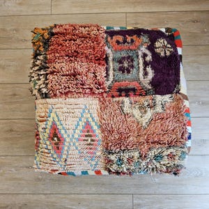 Peut inclure: Un pouf marocain carré multicolore avec un motif patchwork. Le pouf présente un mélange de textures et de couleurs, notamment des nuances de rouge, de rose, de violet et de crème. Le design comprend des motifs géométriques et une texture touffue.