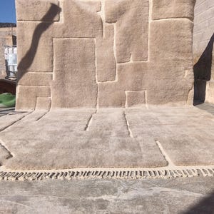 Peut inclure: Un tapis beige avec un motif géométrique, composé de formes rectangulaires et irrégulières. Le tapis est présenté à l'extérieur, avec un ciel bleu clair en arrière-plan. Le tapis a une surface texturée et une frange sur le bord inférieur.