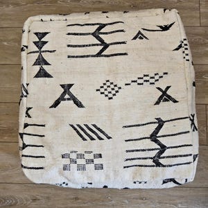 Peut inclure: Pouf carré blanc cassé avec un motif géométrique noir. Le design présente diverses formes, notamment des triangles, des lignes et des figures en forme de croix. Le pouf est posé sur un sol en bois. Le texte "Tifinagh Shop" est visible en bas à gauche.