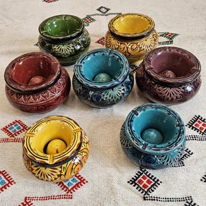 Peut inclure: Ensemble de sept cendriers en céramique de différentes couleurs, dont vert, jaune, turquoise et bordeaux. Chaque cendrier a une forme ronde avec une indentation centrale et des motifs décoratifs. Ils sont disposés sur un tissu blanc à motifs.