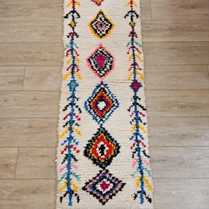 Peut inclure: Un tapis marocain blanc avec des motifs en losange colorés dans des tons de bleu, rouge, jaune, rose et noir. Le tapis a des franges aux extrémités.