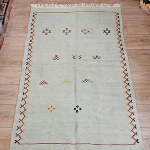 May include: A rectangular, light green rug with a cream-colored fringe. The rug features a repeating geometric pattern in black, red, yellow, and white. The border has a zig-zag design. The text "Tifinagh Shop" is visible.