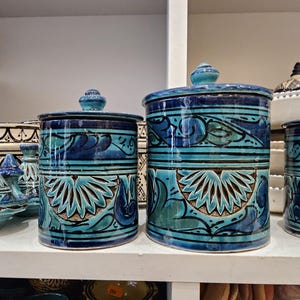 Puede incluir: Dos tarros cilíndricos de cerámica con tapas, adornados con un diseño floral turquesa y azul oscuro. Los tarros se exhiben en una estantería blanca, destacando los intrincados patrones y la artesanía. Los tarros son parte de una colección de cerámica decorativa.