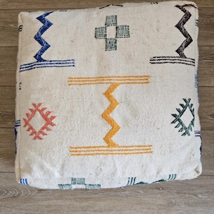 Peut inclure: Pouf carré blanc cassé avec des motifs géométriques colorés. Le design comprend des zigzags, des croix et des losanges bleus, orange, verts et marron. Le pouf est en tissu texturé et repose sur une surface en bois.