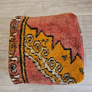 Peut inclure: Un pouf carré texturé aux tons corail, jaune, noir et blanc. Le coussin présente un motif géométrique avec un motif jaune central et des accents noirs. Le logo Tifinagh Shop est visible en bas à gauche.