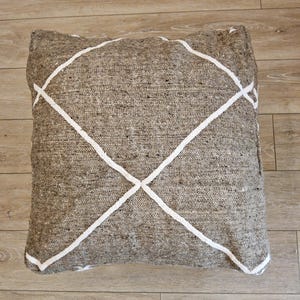 Peut inclure: Coussin de sol carré en tissu marron avec un motif géométrique blanc. Le coussin a un aspect texturé et est posé sur un sol en bois. Le motif est une série de lignes blanches qui s'entrecroisent.