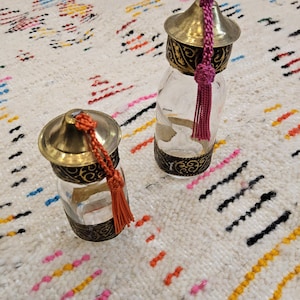 Può includere: Due bottiglie di vetro decorative con tappi in metallo dorato e nero e nappine. Le bottiglie sono su un tappeto bianco e colorato.