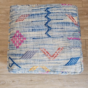 May include: Square blue and white patterned pouf with embroidered geometric designs in pink, red, yellow, and blue. The pouf is made of woven fabric and has a textured appearance. The Tifinagh Shop logo is visible in the bottom left corner.