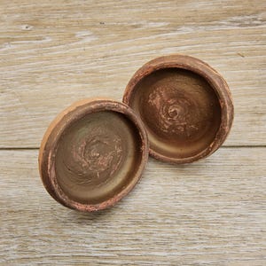 Puede incluir: Dos cuencos pequeños y abiertos de arcilla color terracota con una superficie rugosa y texturizada. Los cuencos son redondos con un borde ligeramente ensanchado y un interior marrón oscuro. Los cuencos están sobre un fondo de vetas de madera clara.