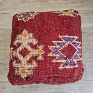Peut inclure: Un pouf marocain carré bordeaux avec des motifs géométriques crème, violets et jaunes. Le pouf est fait d'un tissu texturé et repose sur un sol en bois clair. Le nom de la boutique "Tifinagh Shop" est visible.