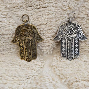 Puede incluir: Dos colgantes de mano de Hamsa, uno dorado y otro plateado, con intrincados diseños de Estrella de David y geométricos. Cada colgante tiene un lazo para colgar. Se muestran sobre una superficie texturizada de color blanco roto.