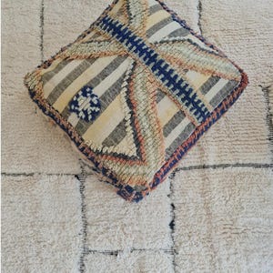 Peut inclure: Un coussin carré de couleur crème avec un motif géométrique dans les tons bleus, roses et gris. Le coussin est fait d'un tissu texturé et a une bordure en frange.