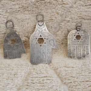 May include: Three silver-colored Hamsa hand pendants with Star of David cutouts. The pendants feature intricate engraved designs and are displayed on a textured, off-white surface. Each pendant has a loop for hanging.