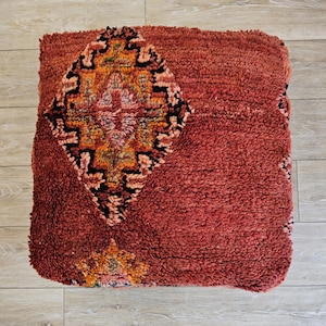 Peut inclure: Un pouf carré rouge texturé avec un motif central en losange noir, orange et rose. Le pouf a un aspect tissé et moelleux, suggérant une assise confortable. Le design rappelle les textiles marocains traditionnels.
