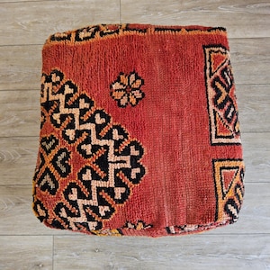 Peut inclure: Un pouf carré rouge avec des motifs géométriques complexes en noir, orange et crème. Le pouf a une surface texturée et un motif floral central. Le logo Tifinagh Shop est visible en bas à gauche.