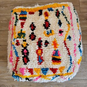 Peut inclure: Un pouf blanc et moelleux avec un motif géométrique coloré. Le motif comprend du rouge, du jaune, du bleu, du vert et du noir. Le pouf est de style marocain traditionnel.