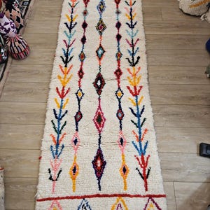 Peut inclure: Un tapis marocain blanc avec un motif géométrique coloré. Le tapis a une frange en bas et est fait de laine.