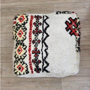 Peut inclure: Un coussin de sol carré blanc avec un motif géométrique noir, rouge et jaune. Le coussin est fait d'un matériau doux et moelleux.