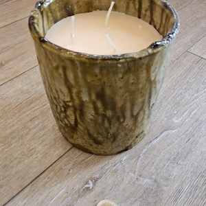 May include: A large, cylindrical candle in a rustic, glazed ceramic container. The candle has three wicks and a creamy, off-white wax. The container is a mottled brown and tan colour, with a textured surface. A two-euro coin is next to the candle.