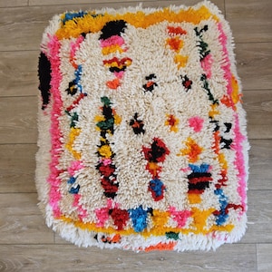 Peut inclure: Un pouf blanc et moelleux avec un motif géométrique coloré dans des tons de rose, jaune, orange, bleu, vert et noir. Le pouf est en laine et a une bordure en franges.