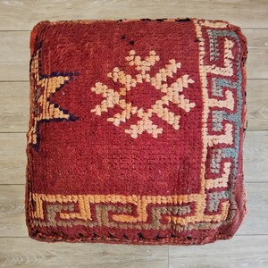 Peut inclure: Un pouf marocain carré rouge avec des motifs géométriques beiges, oranges et bleus. Le pouf a un aspect tissé texturé et est placé sur un sol en bois clair. Le pouf est un objet décoratif.