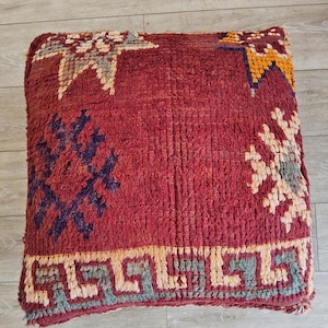 Peut inclure: Un pouf carré rouge texturé avec des motifs géométriques blancs, jaunes et bleus. Le pouf présente un motif de bordure et des formes en étoile. Le logo Tifinagh Shop est visible en bas à gauche.