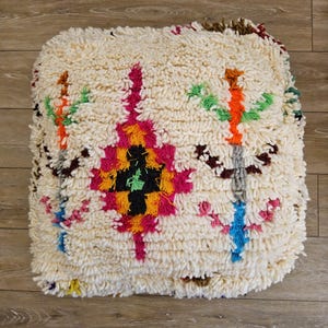 Peut inclure: Un coussin de sol carré en laine à poils longs de couleur crème, orné d'un motif géométrique central en losange aux couleurs vives magenta, orange et noir, encadré par deux motifs floraux abstraits verticaux. Le texte Tifinagh Shop est visible.